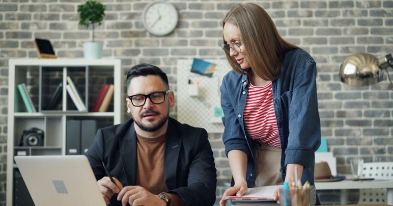 A woman hovering over a man looking at a laptop at work