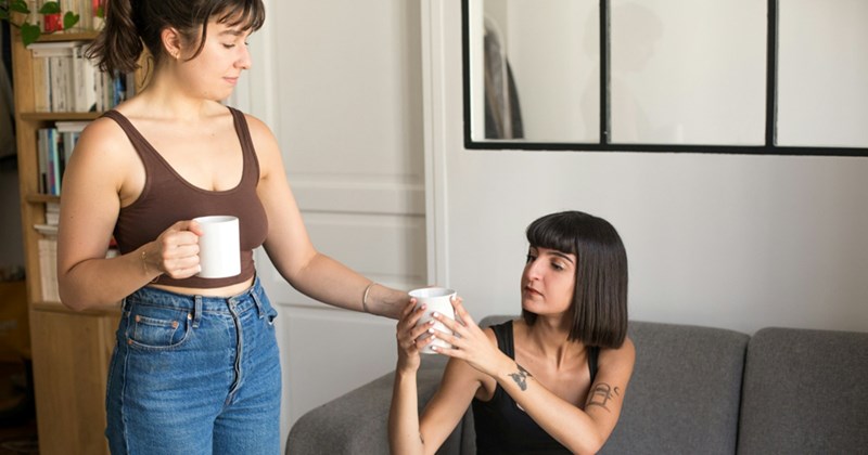 A woman who is standing holds two mugs, passing one to a woman sat on a couch beside her