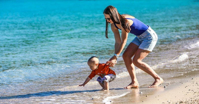 a woman wearing a bathing suit and shorts leans down and holds the hand of a toddler who is crouching at the edge of the sea surf