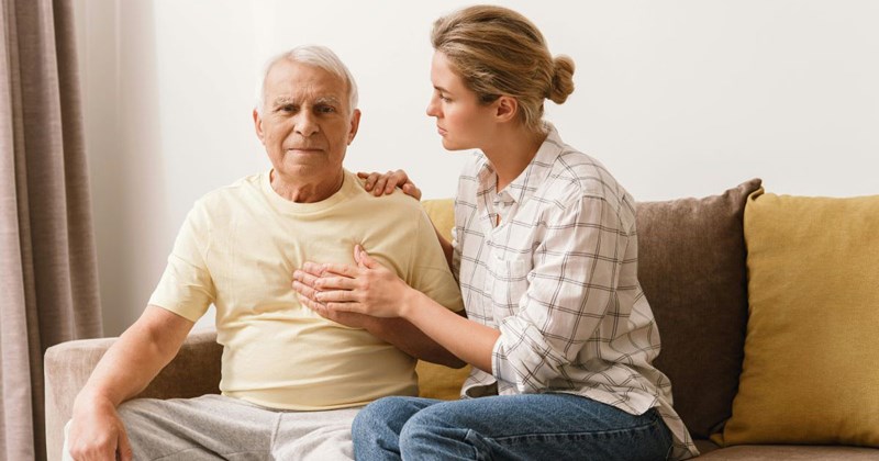 Anxious woman sitting with her father-in-law on the couch at his house, laying her hand on his hand as he clutches his chest