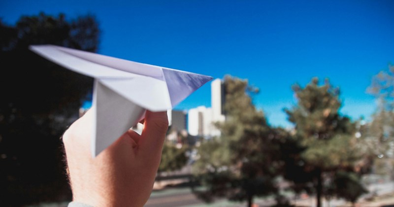 A hand holds a paper plane as if to throw it, with blue sky, trees, and high rise buildings in the background