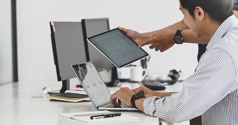 An employee is seen from the side, typing on a laptop. In the background, a manager holds a tablet to discuss a project or ticket.