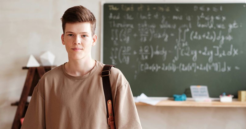Teenage boy standing in front of a classroom chalkboard and looking smug at the camera