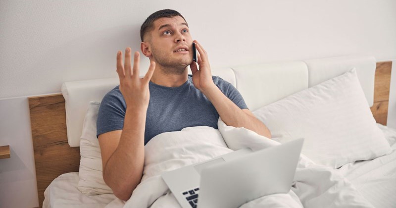 Unemployed man lies in bed while looking on his laptop and talking on the phone