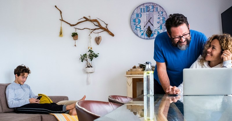 a man stands over a woman who is sat by a laptop and looking up at him smiling while a teenager looks at his phone while sat on the sofa in the background